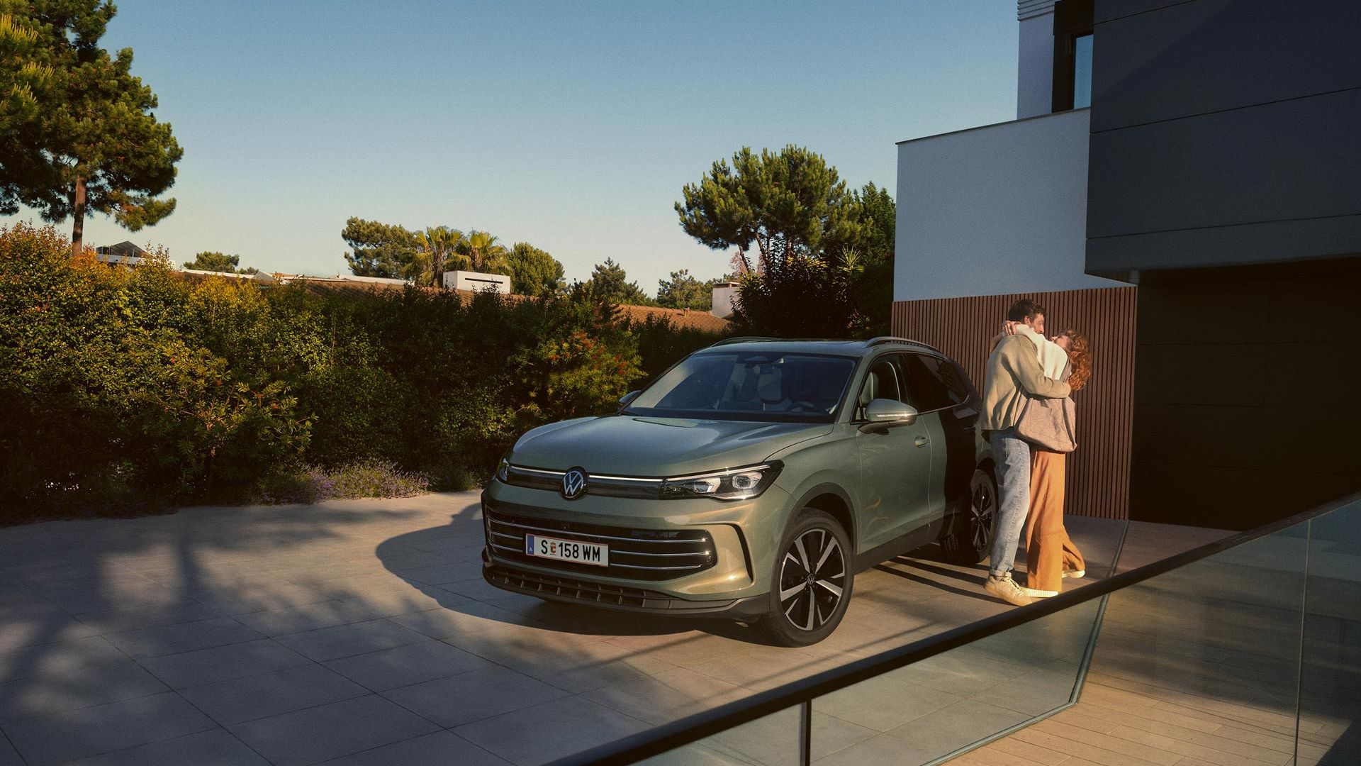 Eine Frau und ein Mann umarmen sich, neben ihnen steht ein VW Tiguan Elegance in der Farbe Cipressino-Grün Metallic