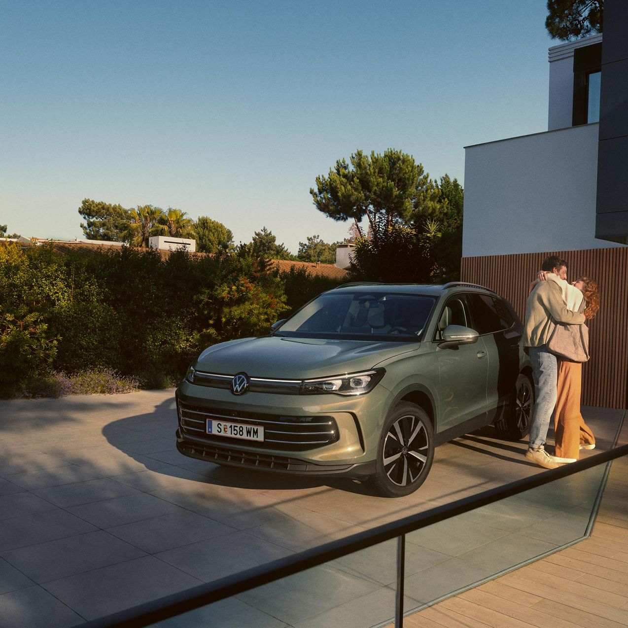 Eine Frau und ein Mann umarmen sich, neben ihnen steht ein VW Tiguan Elegance in der Farbe Cipressino-Grün Metallic