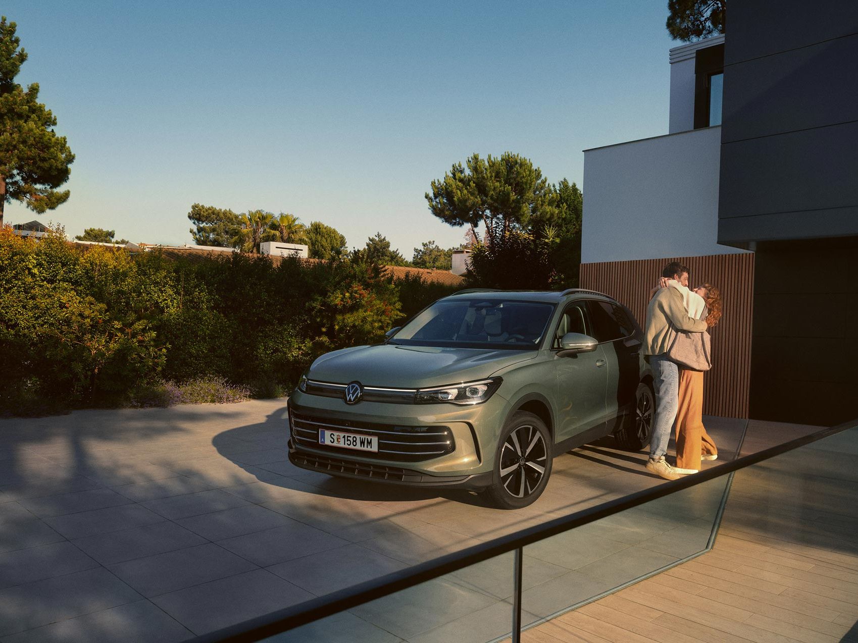 Eine Frau und ein Mann umarmen sich, neben ihnen steht ein VW Tiguan Elegance in der Farbe Cipressino-Grün Metallic