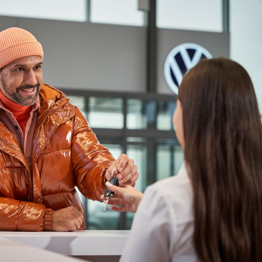 Ein Mann in orangener Jacke gibt Autoschlüssel beim Händler ab