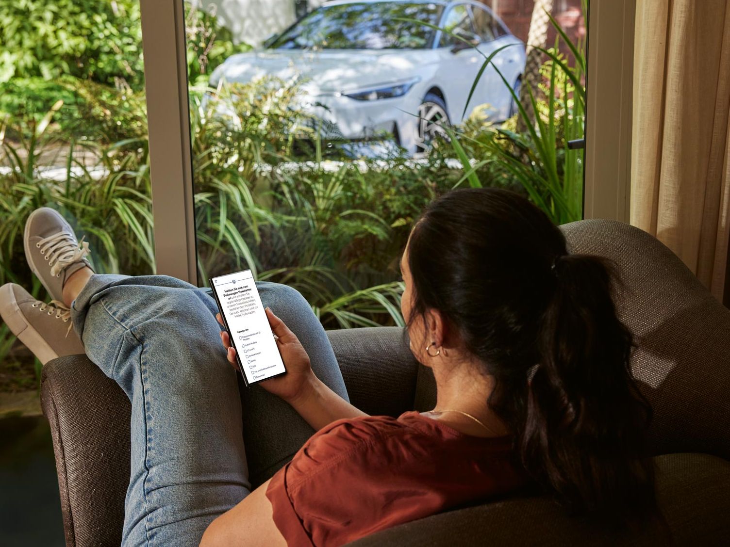 Eine Frau sitzt entspannt im Sofa und blickt auf ihr Smartphone, im Hintergrund ein VW ID.7