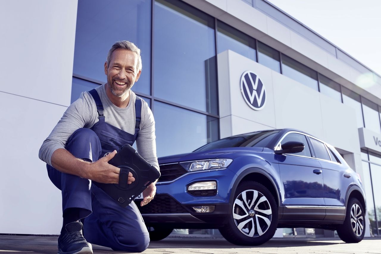 Ein Volkswagen Service Techniker mit Tablet, kniet vor einem blauen Volkswagen T-Roc