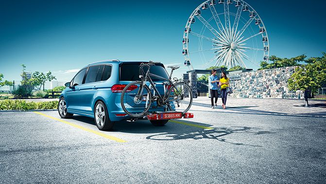 Ein VW Touran in blau mit Fahrradträger parkt auf einem Parkplatz, im Hintergrund steht ein Riesenrad.