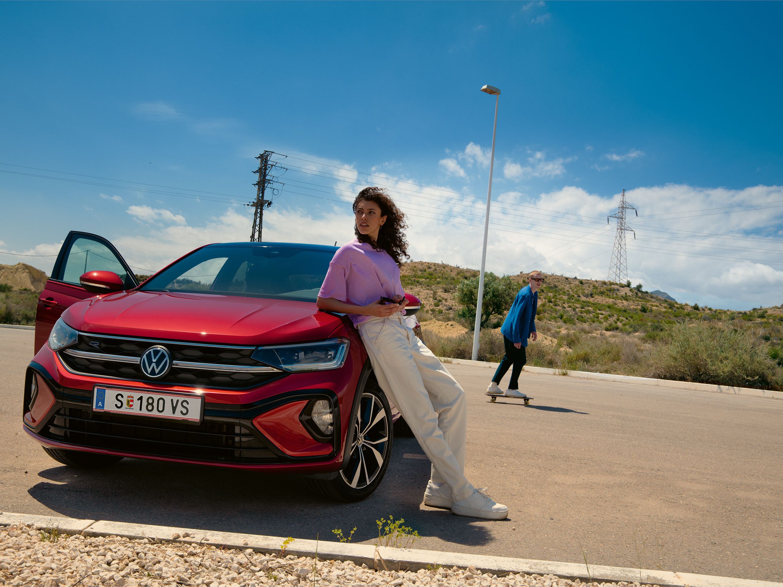 VW Taigo R-Line in Rot, Frontansicht auf Parkplatz. Frau lehnt an Motorhaube, Mann fährt Skateboard