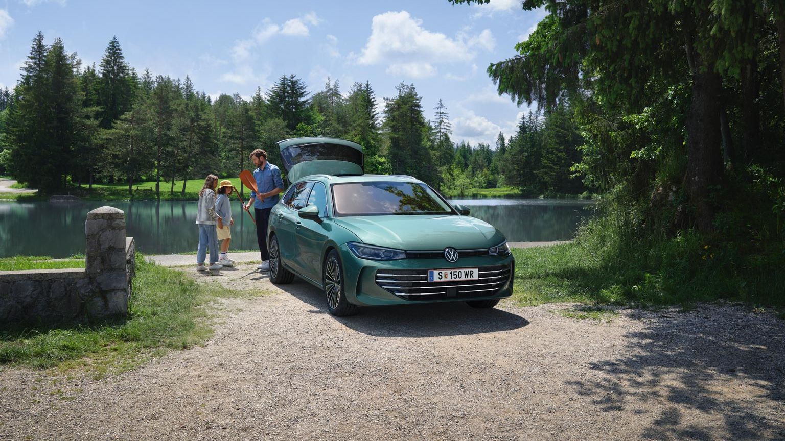 Der neue VW Passat Elegance in Maripositgrün Metallic parkt an einem Badesee mit geöffnetem Kofferraum. Ein Mann verteilt Freitzeitspielzeug an zwei Kinder.