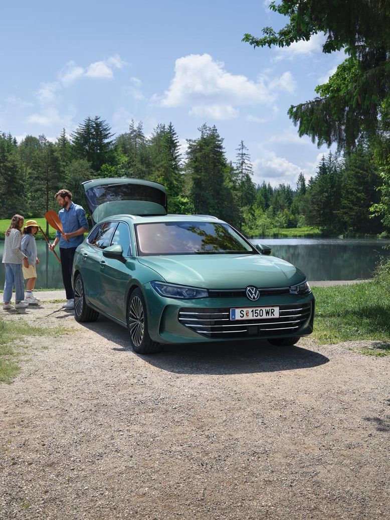 Der neue VW Passat Elegance in Maripositgrün Metallic parkt an einem Badesee mit geöffnetem Kofferraum. Ein Mann verteilt Freitzeitspielzeug an zwei Kinder.