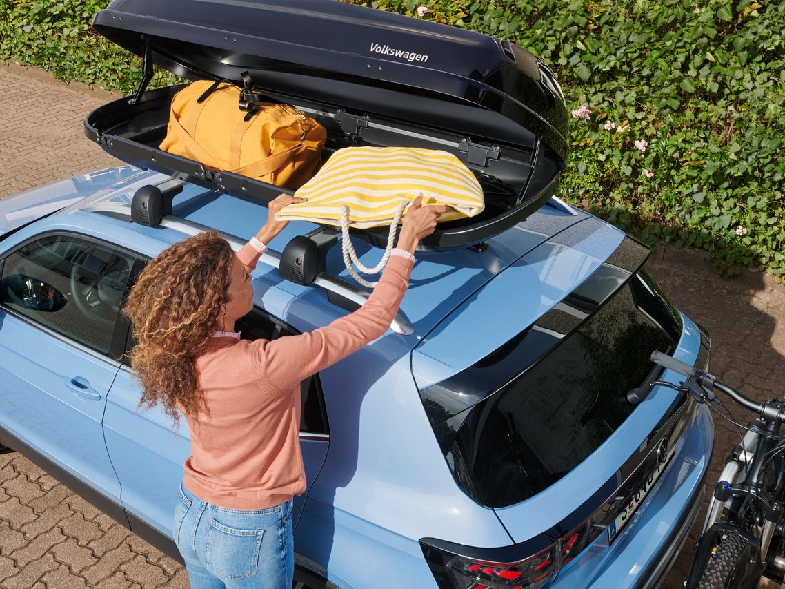 Eine Frau belädt eine Dachbox, die auf dem Dach eines hellblauen Volkswagen T-Cross montiert ist