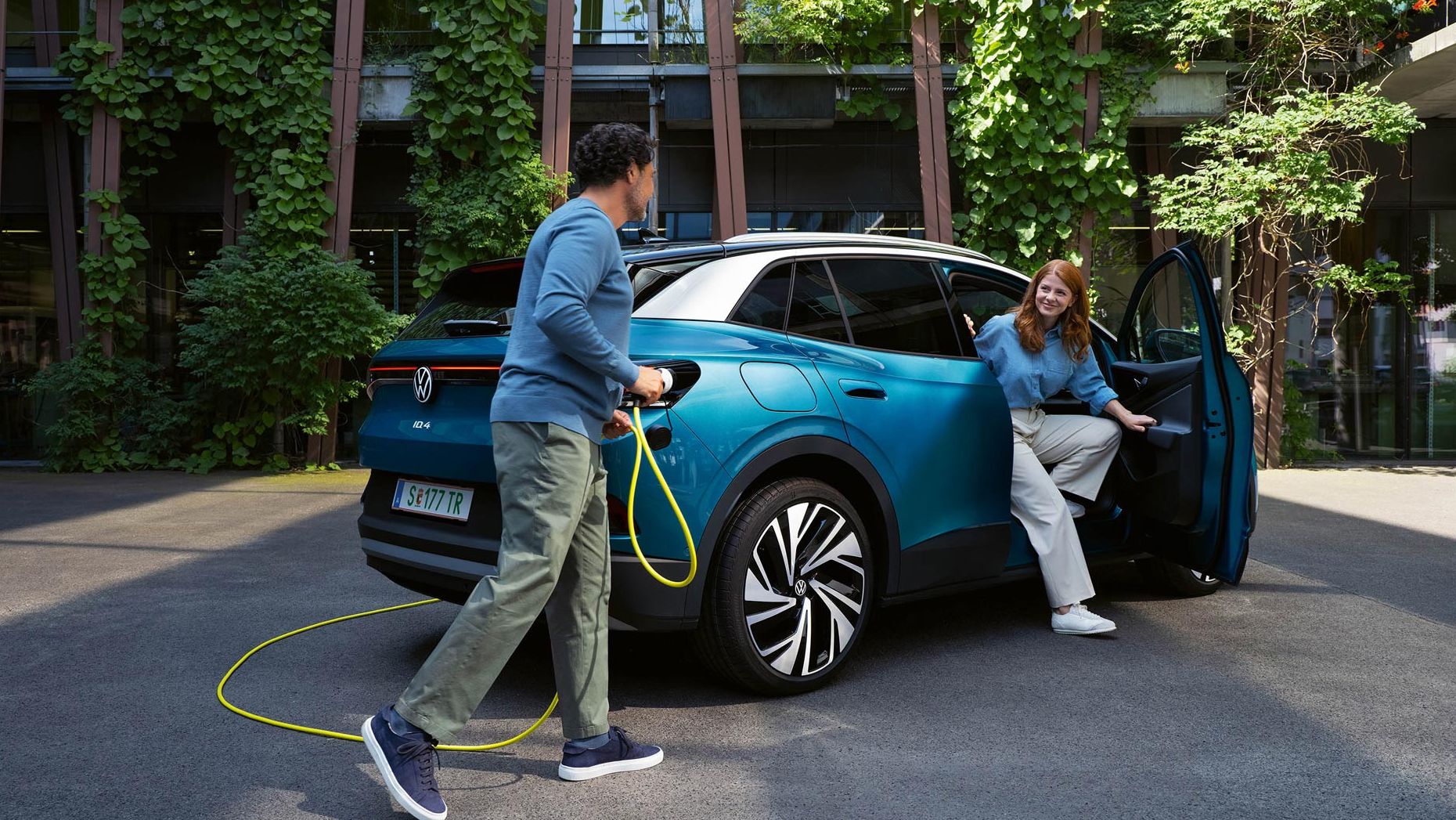 Ein Mann mit einem gelben Ladekabel für Elektroautos geht auf das Heck eines parkenden blauen VW ID.4 zu. Aus der Beifahrertür steigt eine junge Frau aus.