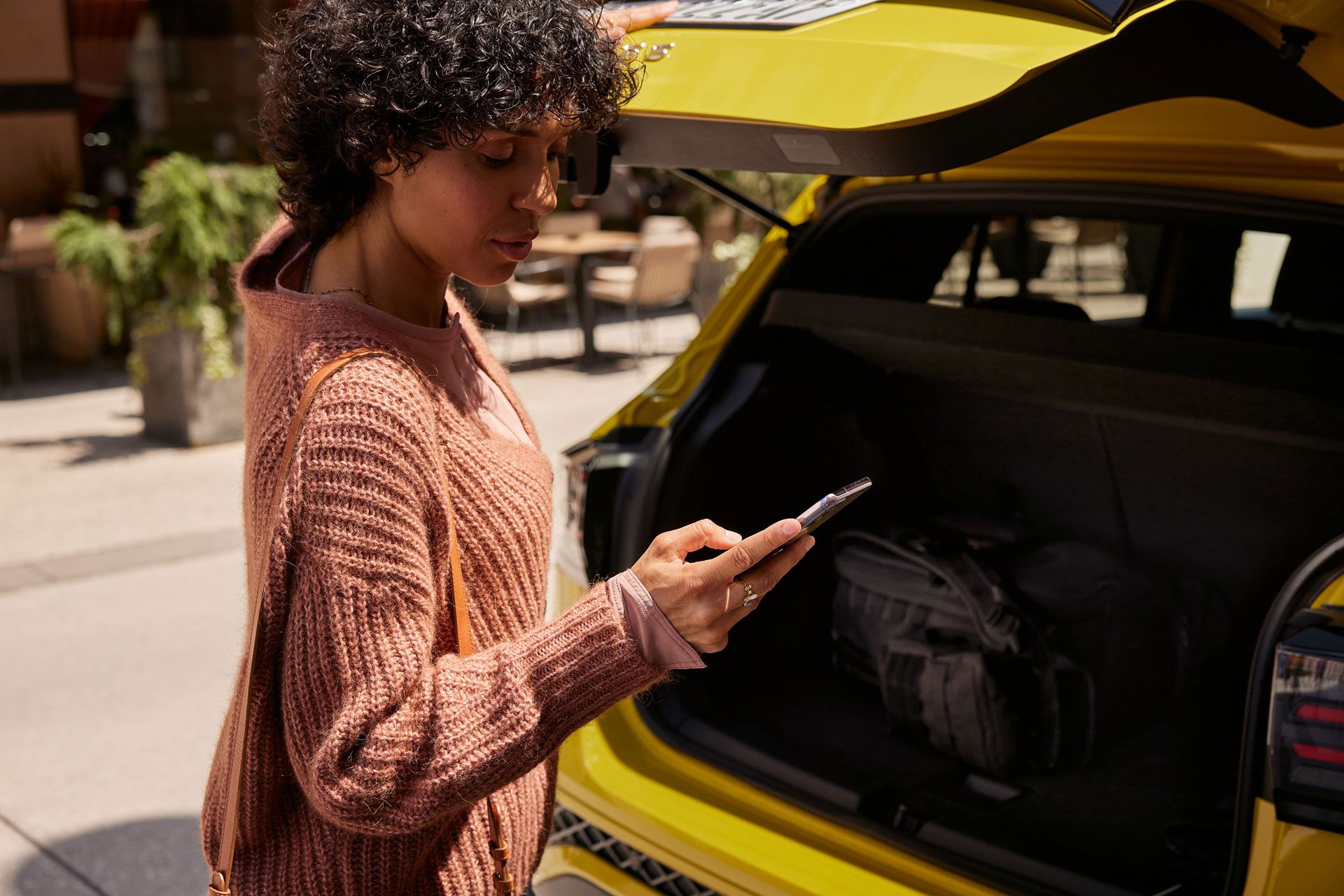 Eine frau schließt den Kofferraum eines gelben VW T-Cross und blickt dabei auf ihr Smartphone. 