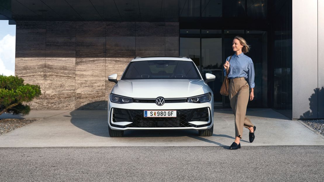 Frontansicht eines weißen VW Passat R-Line, eine Frau geht an ihm vorbei.