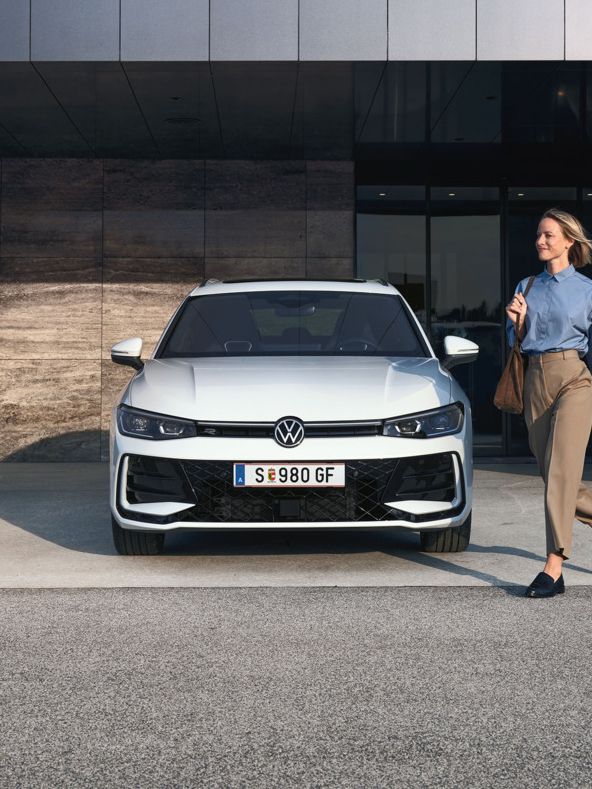 Frontansicht eines weißen VW Passat R-Line, eine Frau geht an ihm vorbei.