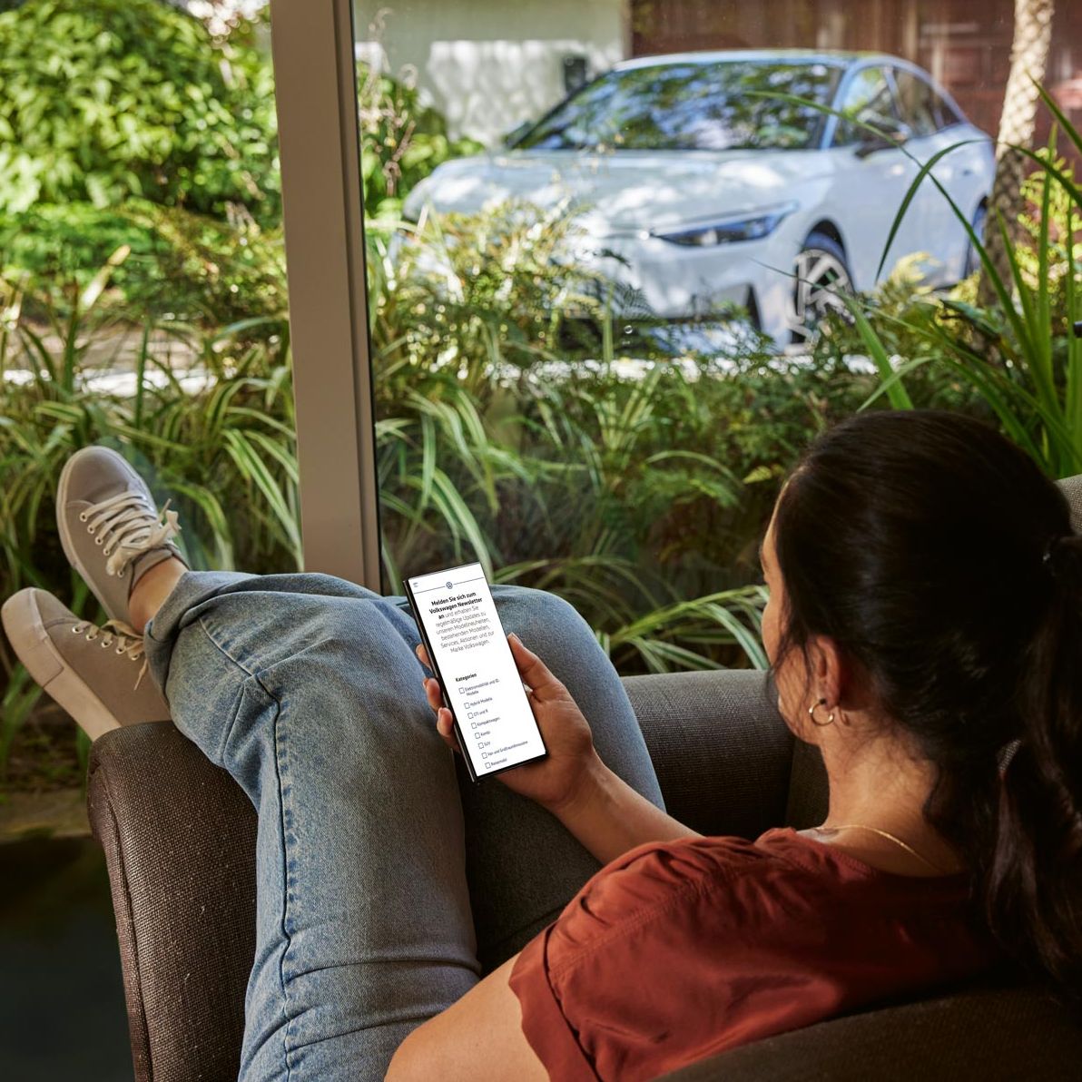 Eine Frau sitzt entspannt im Sofa und blickt auf ihr Smartphone, im Hintergrund ein VW ID.7