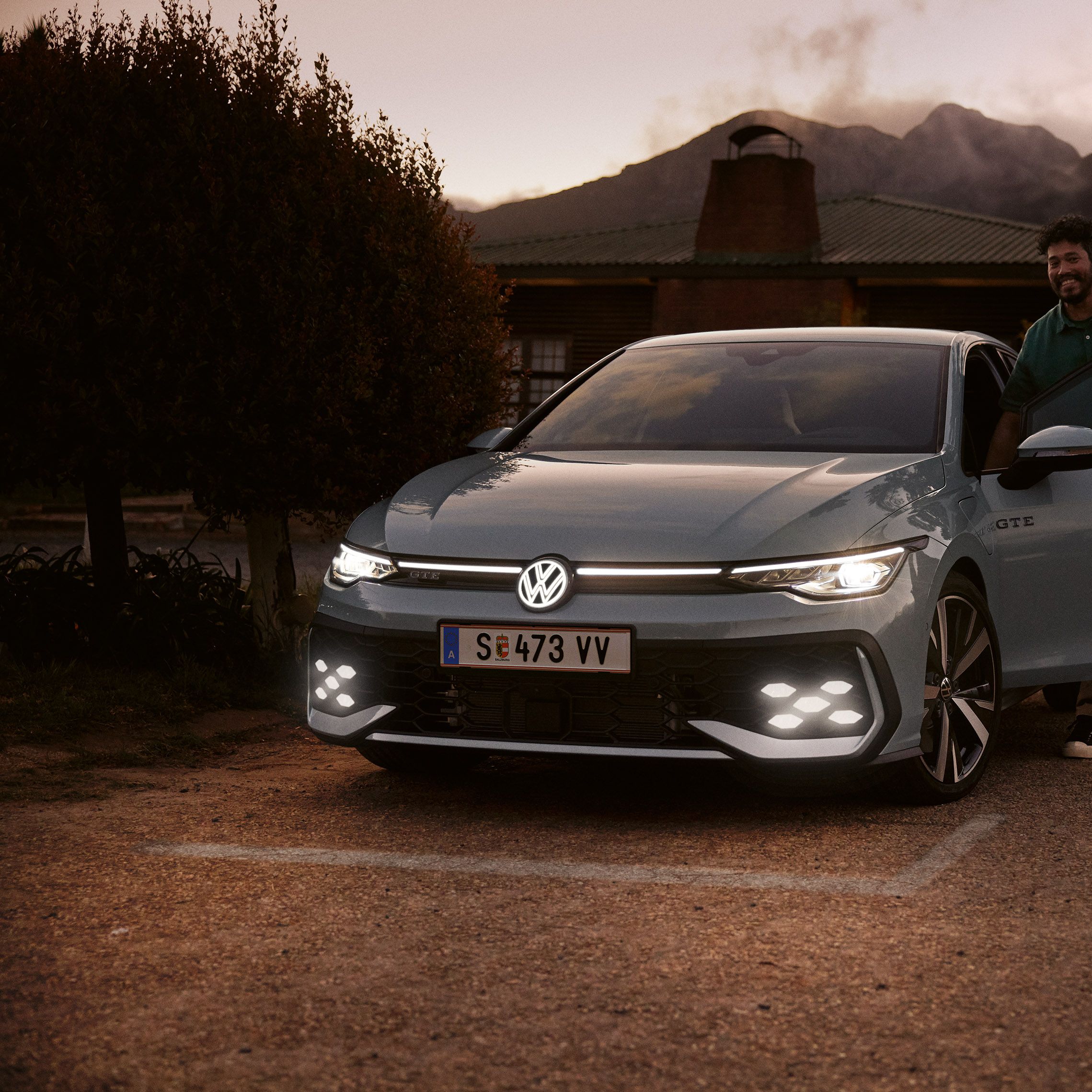 Frontansicht eines grauen VW Golf GTE mit IQ. Light LED Matrix Scheinwerfern in abendlicher Stimmung, Mann steigt in Golf GTE