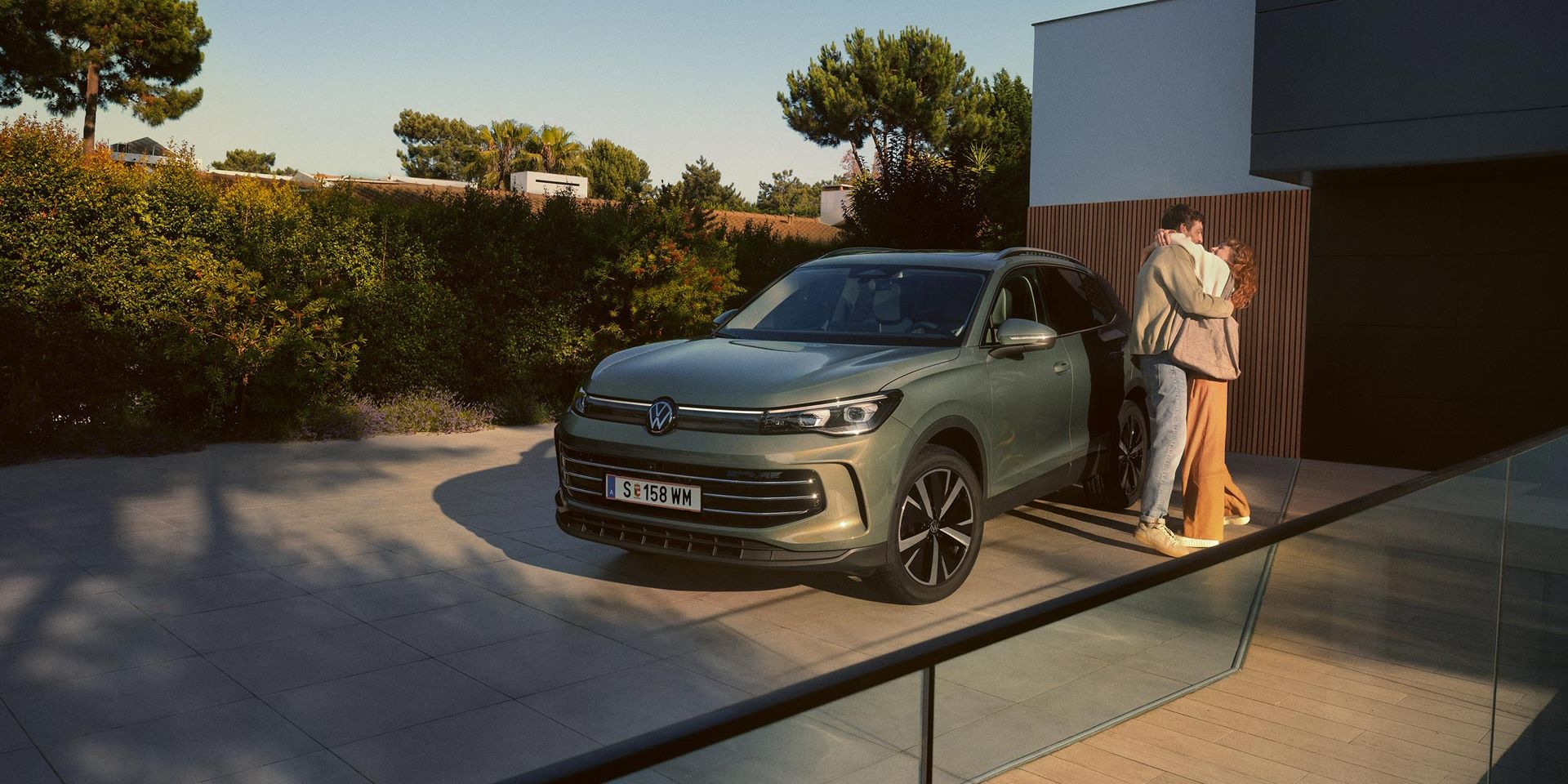 Eine Frau und ein Mann umarmen sich, neben ihnen steht ein VW Tiguan Elegance in der Farbe Cipressino-Grün Metallic