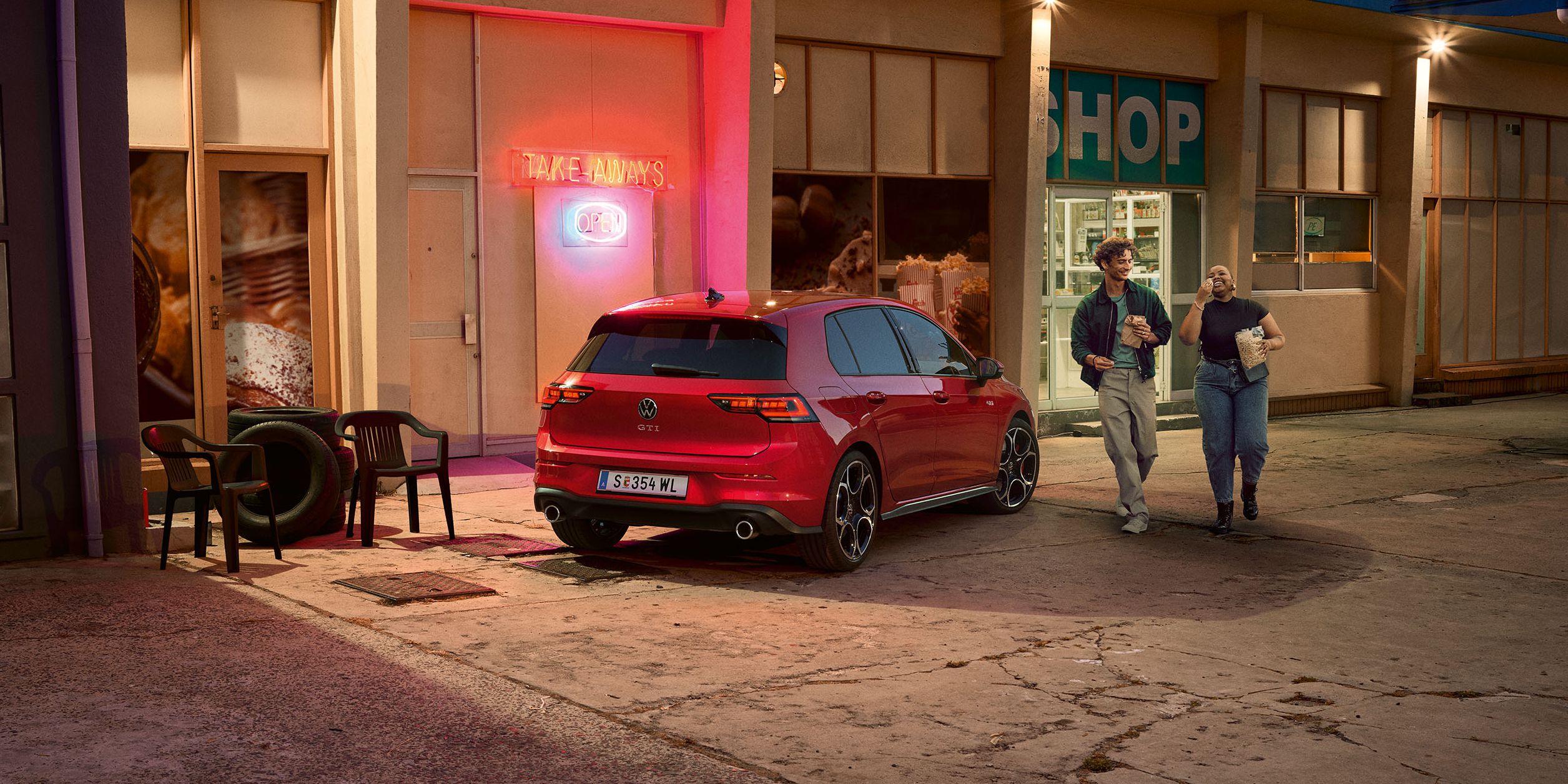 Heckansicht eines roten VW Golf GTI in abendlicher Stimmung, zwei Personen gehen neben ihm her