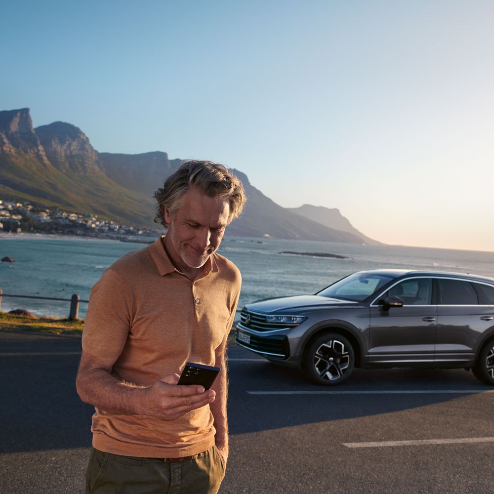 Ein Mann schaut auf sein Smartphone, dahinter parkt ein VW Touareg Elegance, im Hintergrund das Meer und die Küste.