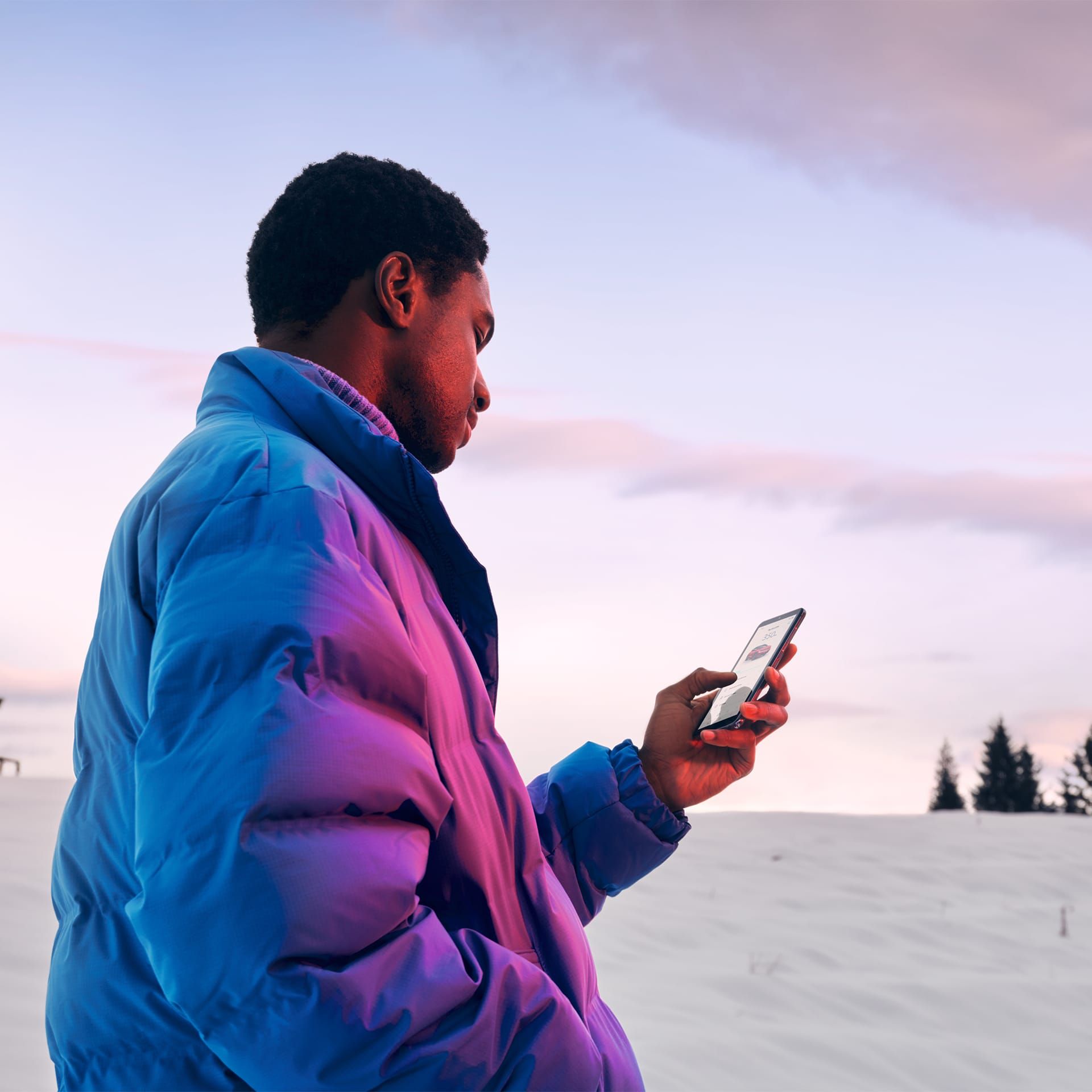 Ein Mann in Winterjacke steht in verschneiter Landschaft und schaut auf sein Smartphone.