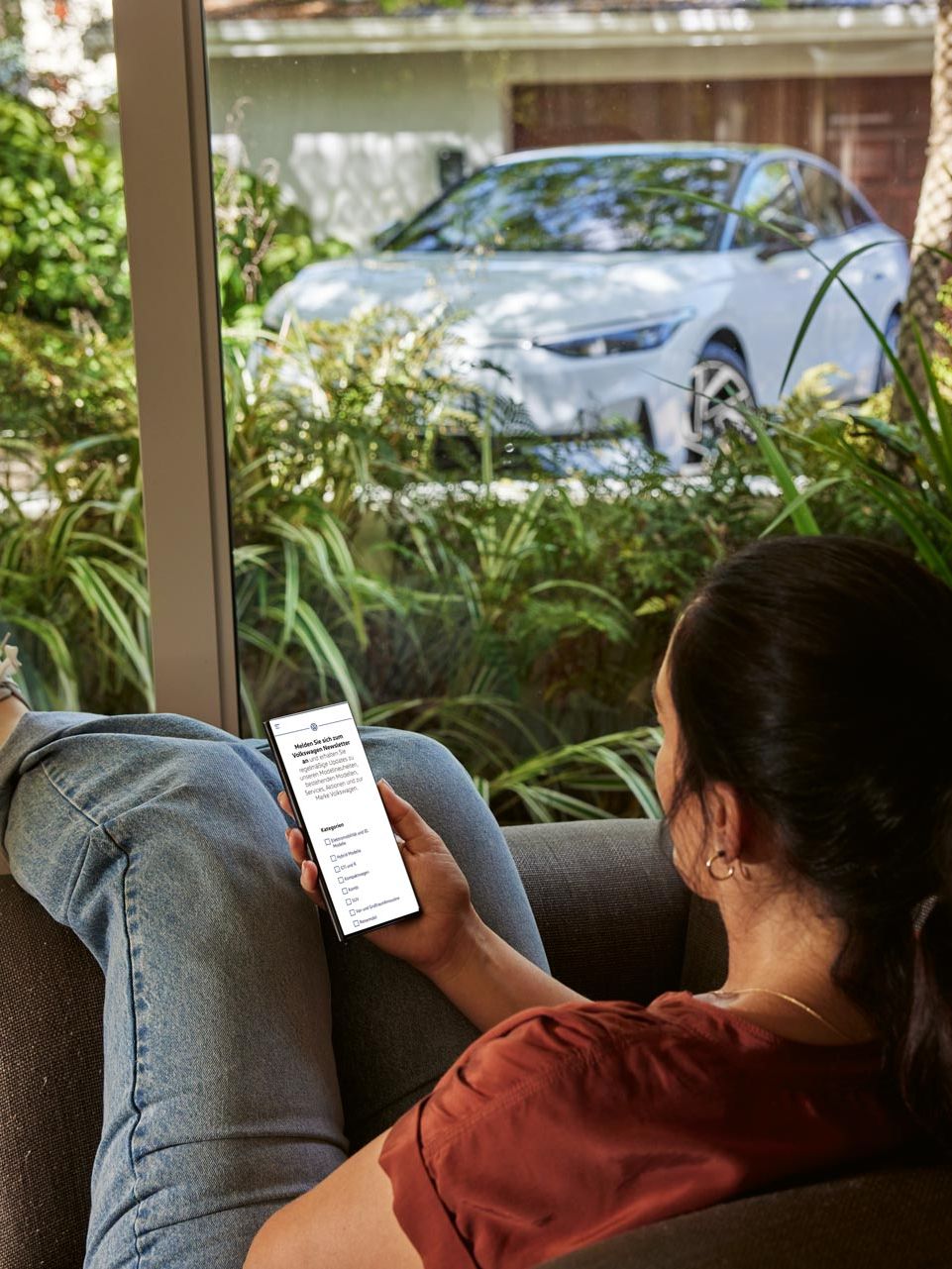 Eine Frau sitzt entspannt im Sofa und blickt auf ihr Smartphone, im Hintergrund ein VW ID.7