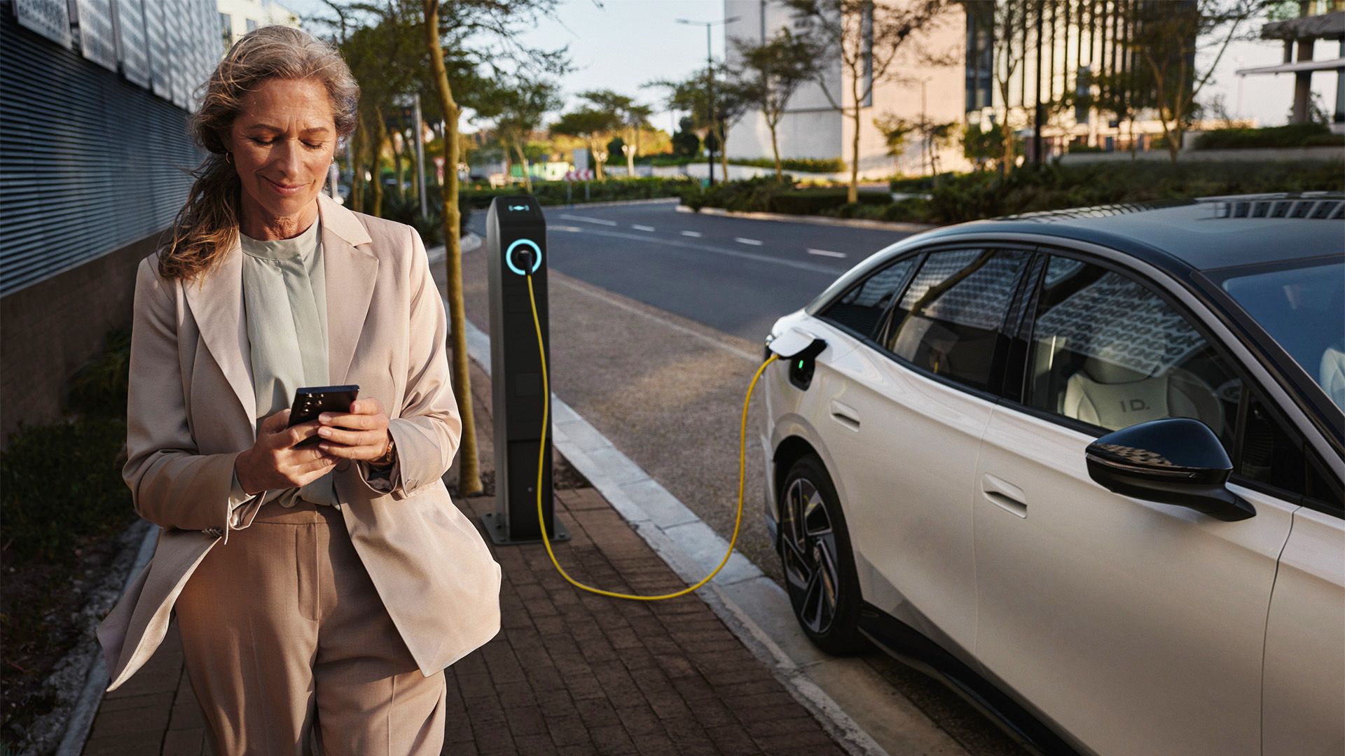 Weißer VW ID.7 seitlich lädt an einer öffentlichen Ladesäule. Eine Frau geht mit dem Smartphone in der Hand daran vorbei.