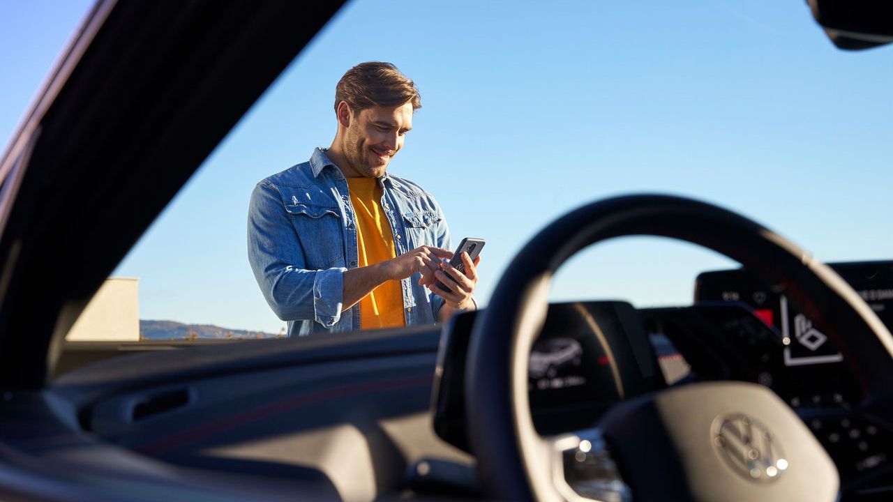 Blick aufs Cockpit und durch die Windschutzscheibe eines Volkswagen. Ein Mann davor tippt in sein Smartphone.