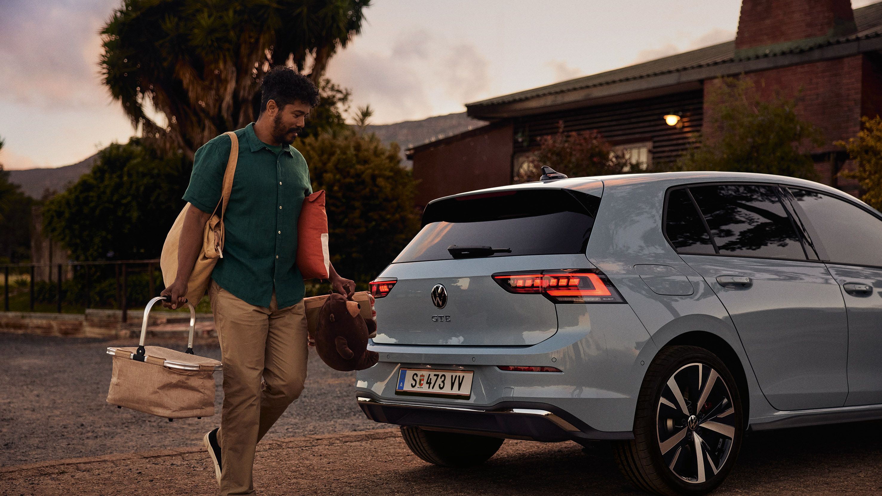 Heckansicht eines grau-blauen VW Golf GTE in abendlicher Stimmung, Mann trägt einen Korb und Spielsachen