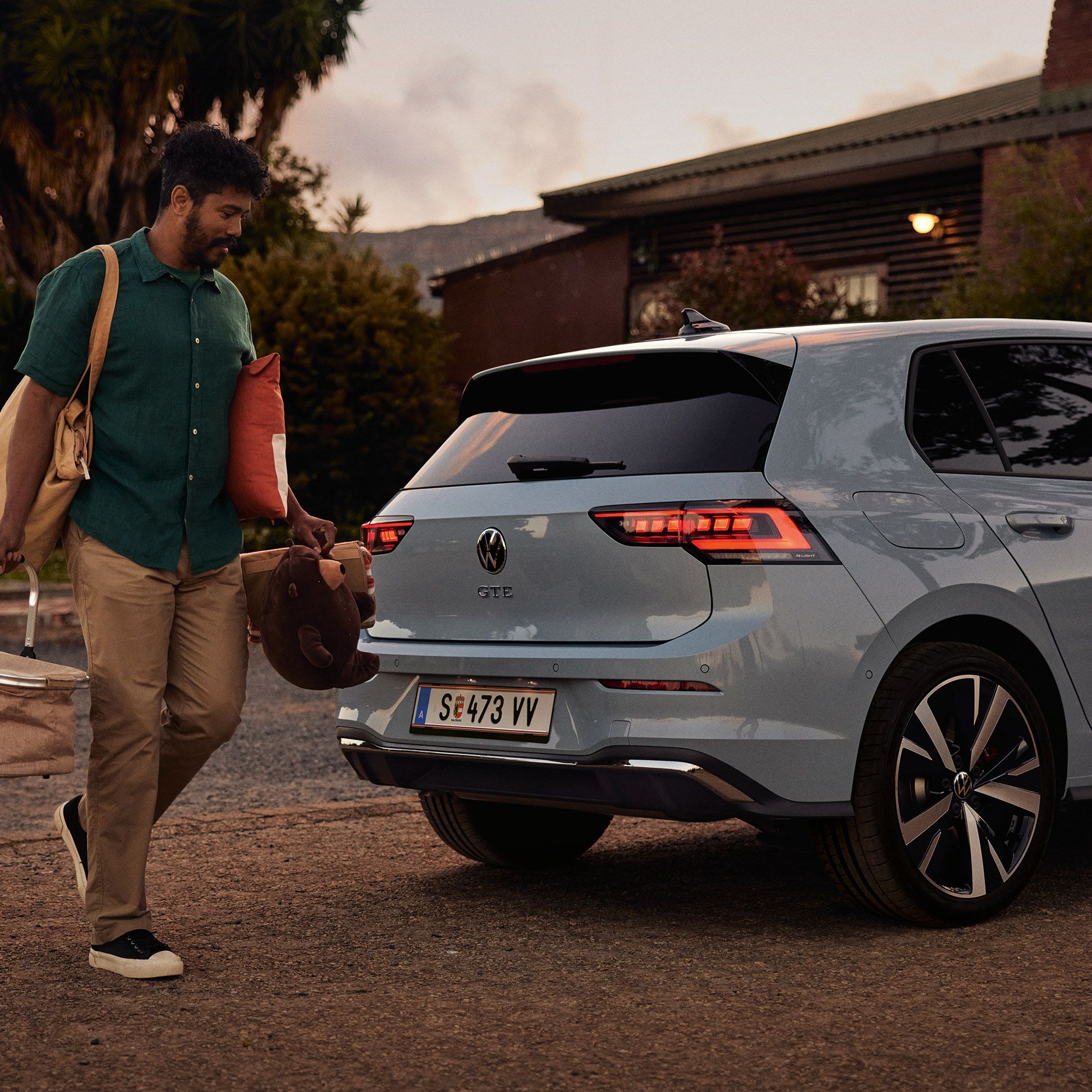 Heckansicht eines grau-blauen VW Golf GTE in abendlicher Stimmung, Mann trägt einen Korb und Spielsachen