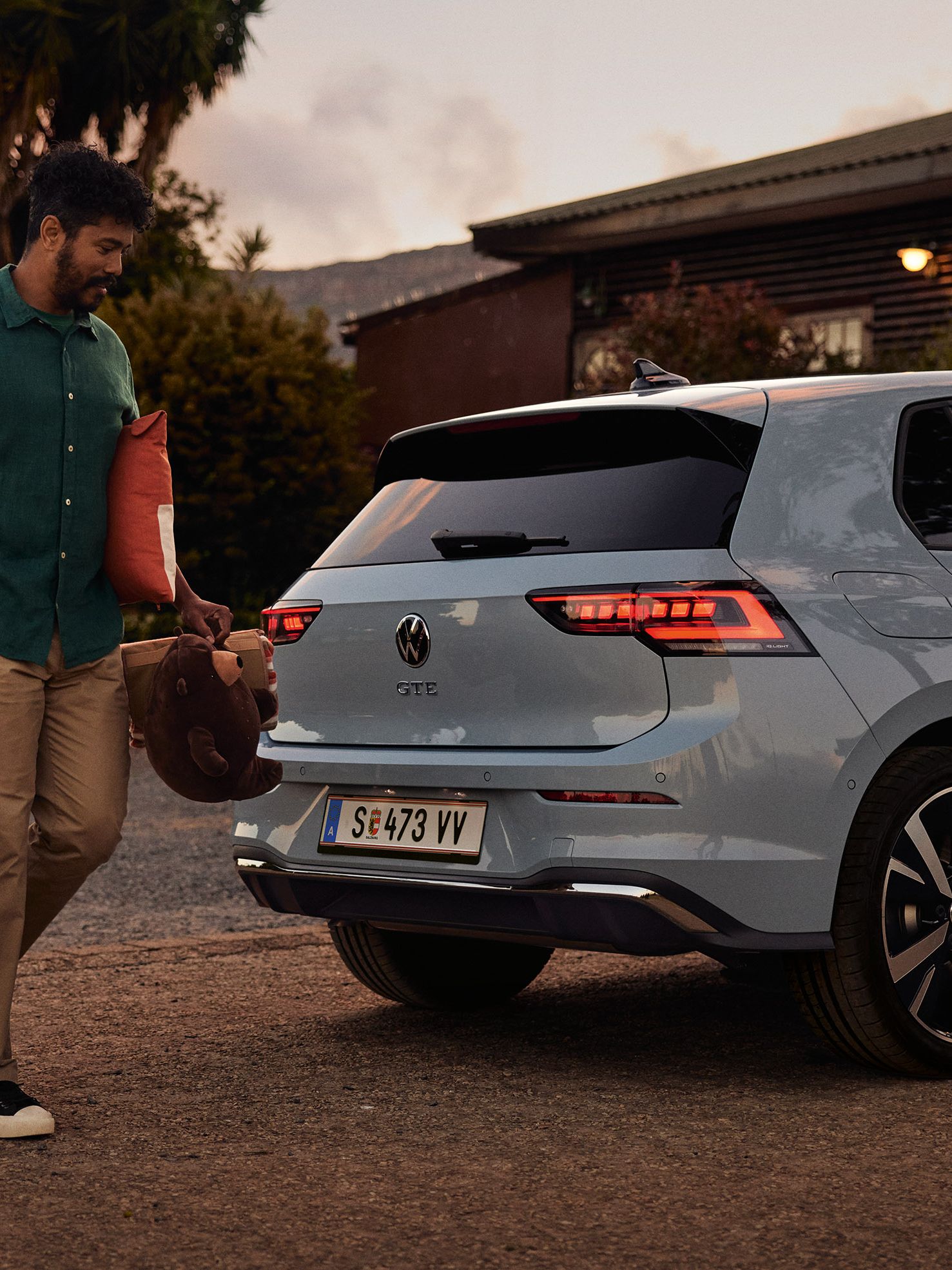 Heckansicht eines grau-blauen VW Golf GTE in abendlicher Stimmung, Mann trägt einen Korb und Spielsachen