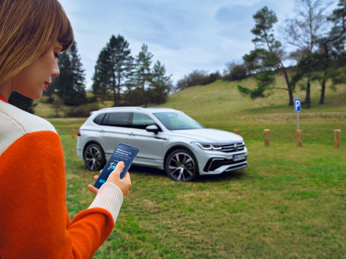 Eine Frau nutzt die Volkswagen App auf ihrem Smartphone, im Hintergrund steht der Tiguan Allspace auf einer Wiese