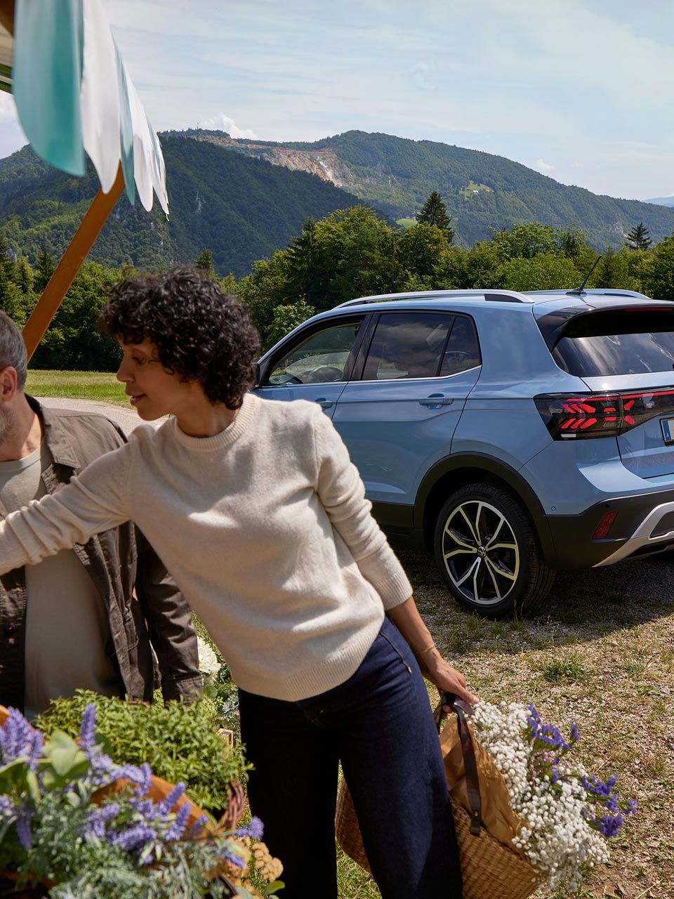 Mann und Frau stehen bei einem Blumenstand, hinter ihnen ein blauer VW T-Cross vor bergiger Landschaft.