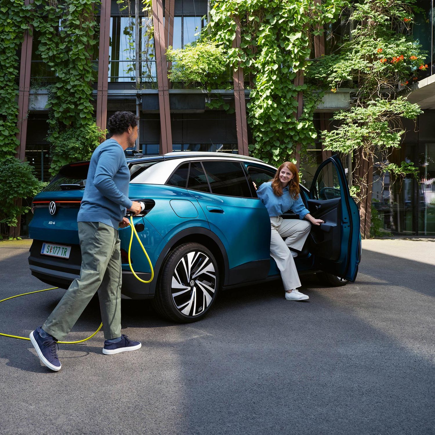 Ein Mann mit einem gelben Ladekabel für Elektroautos geht auf das Heck eines parkenden blauen VW ID.4 zu. Aus der Beifahrertür steigt eine junge Frau aus.