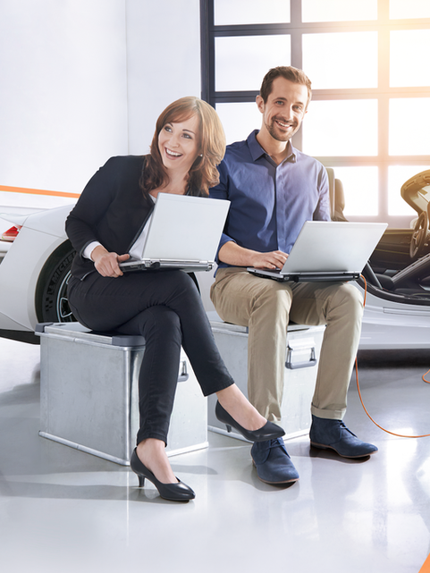 Mann und Frau sitzen mit Laptop vor einem Porsche 