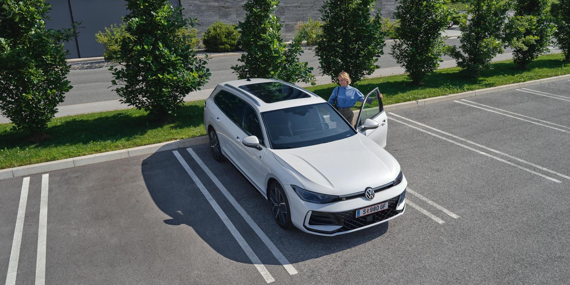 Topshot eines weißen VW Passat R-Line auf einem Parkplatz, eine Frau steigt auf der Fahrerseite ein.