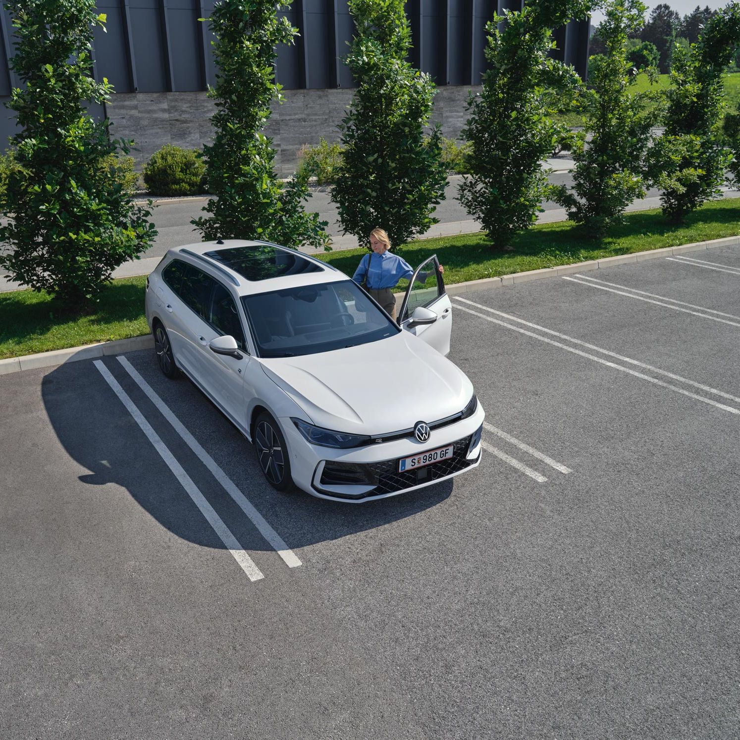 Topshot eines weißen VW Passat R-Line auf einem Parkplatz, eine Frau steigt auf der Fahrerseite ein.