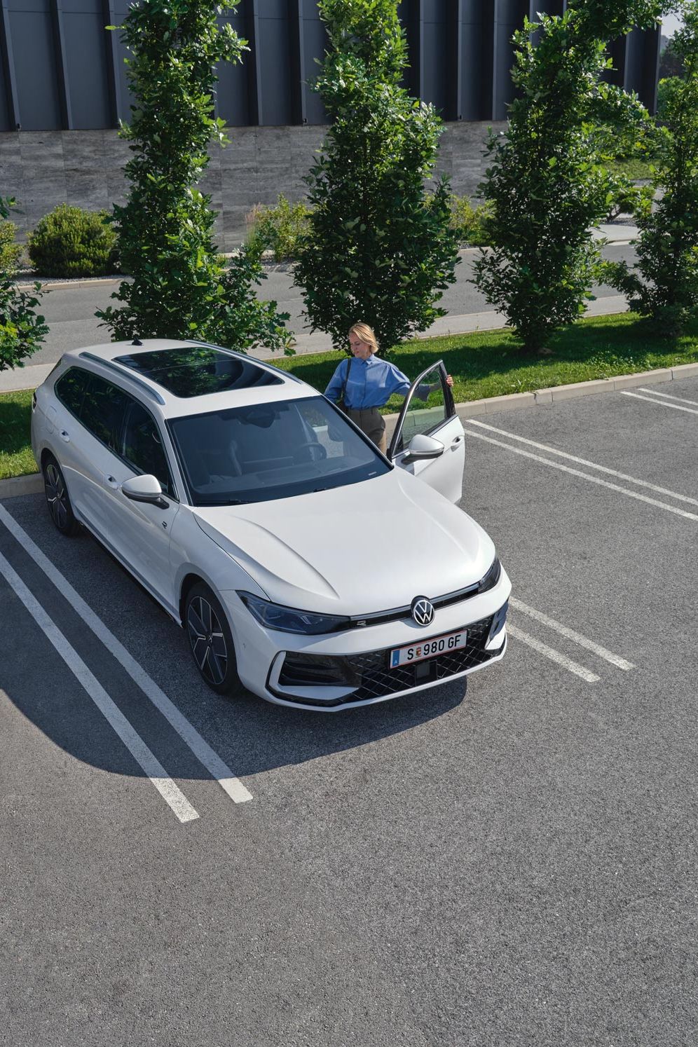 Topshot eines weißen VW Passat R-Line auf einem Parkplatz, eine Frau steigt auf der Fahrerseite ein.