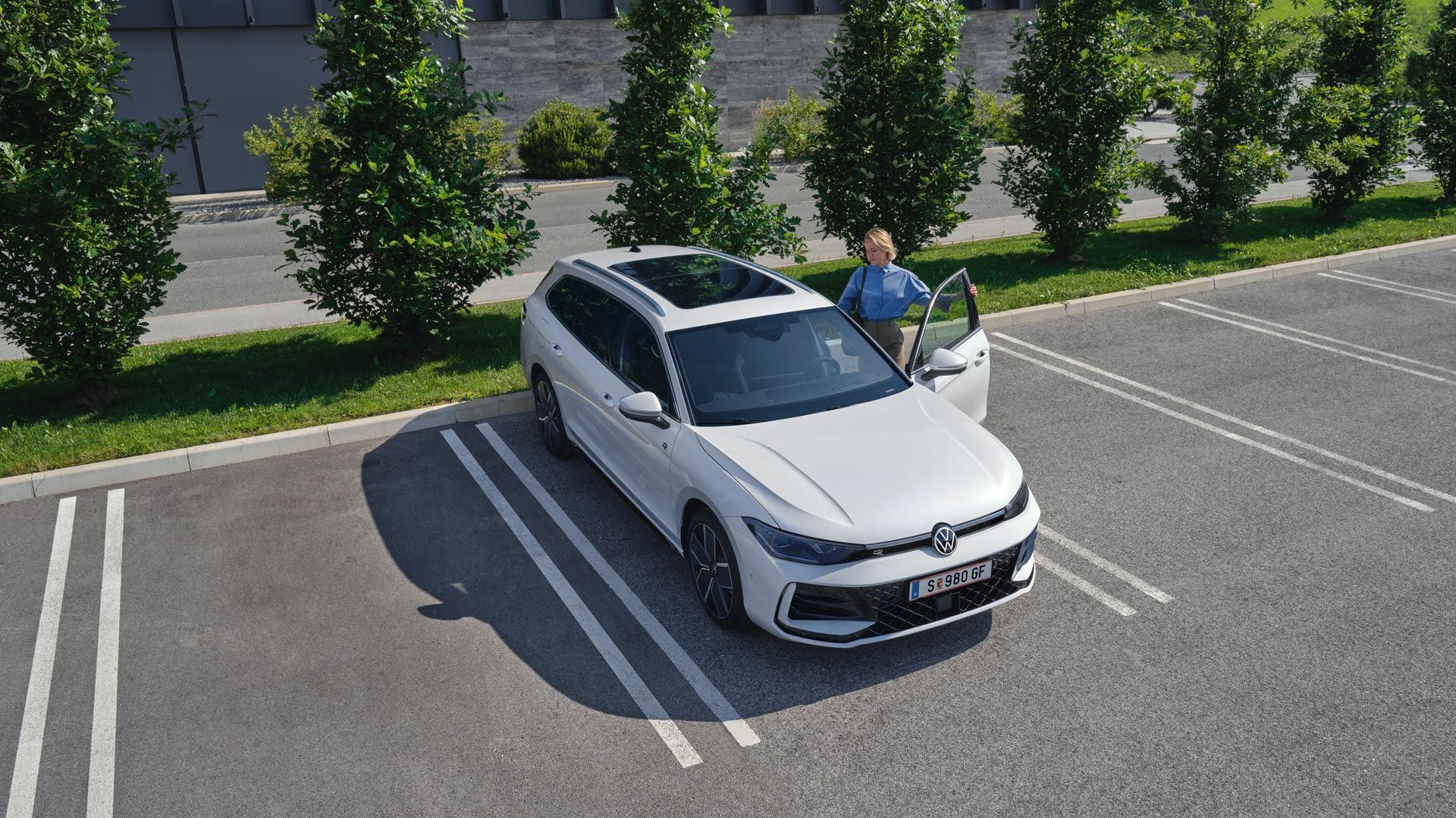 Topshot eines weißen VW Passat R-Line auf einem Parkplatz, eine Frau steigt auf der Fahrerseite ein.