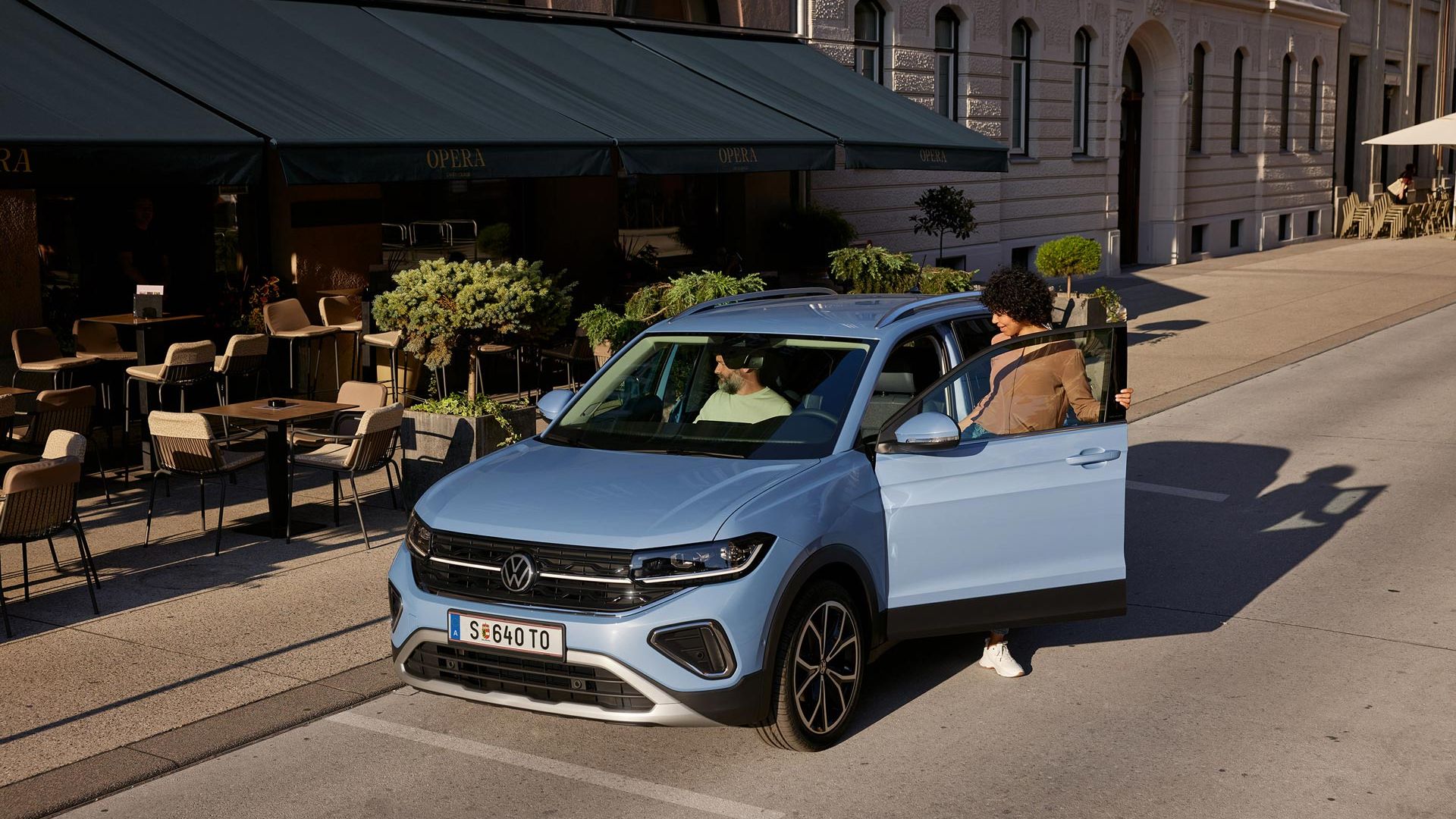 Eine Frau steigt auf der Fahrerseite in den blauen VW T-Cross Style, der vor einem urbanen Straßencafé parkt.