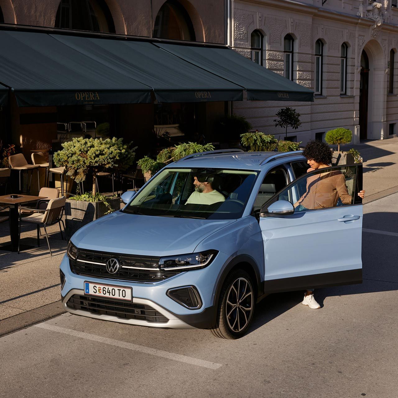 Eine Frau steigt auf der Fahrerseite in den blauen VW T-Cross Style, der vor einem urbanen Straßencafé parkt.