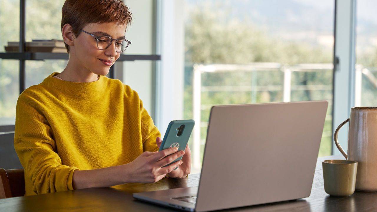 Frau schaut vor ihrem Laptop auf Ihr Handy und prüft, wo ihr Volkswagen gerade ist.