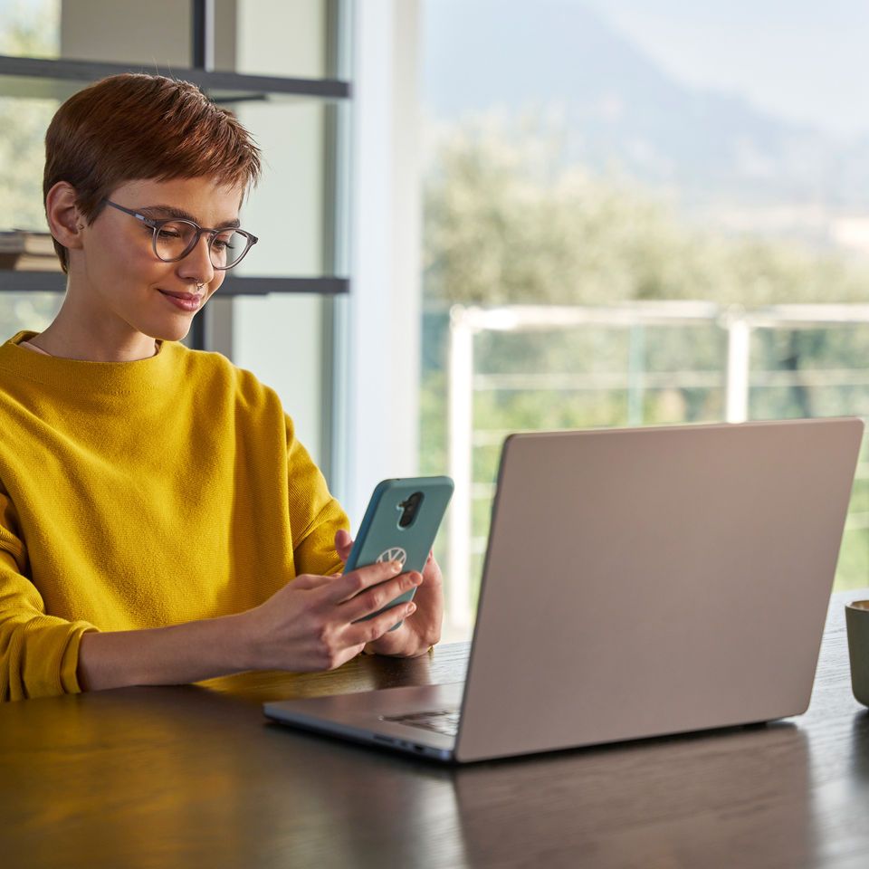 Frau schaut vor ihrem Laptop auf Ihr Handy und prüft, wo ihr Volkswagen gerade ist.