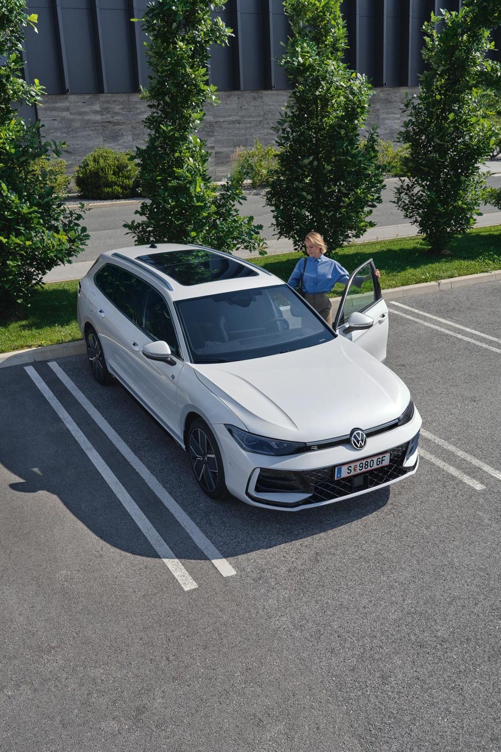 Topshot eines weißen VW Passat R-Line auf einem Parkplatz, eine Frau steigt auf der Fahrerseite ein.