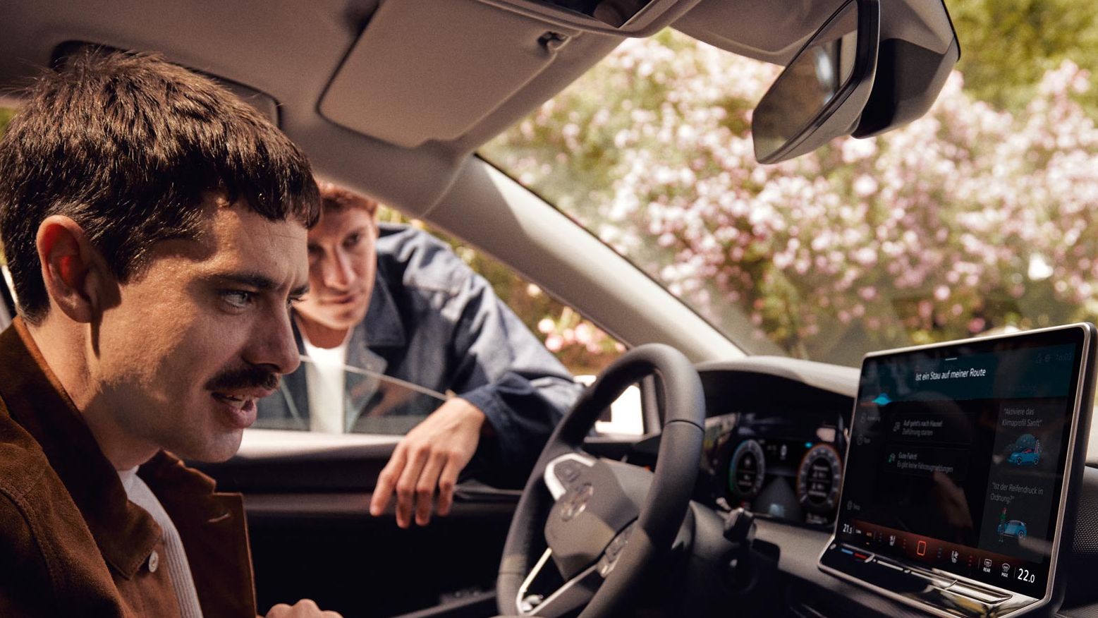 Ein Mann sitzt im neuen VW Golf und schaut auf das Infotainment-Display, ein anderer schaut durch das Fenster.