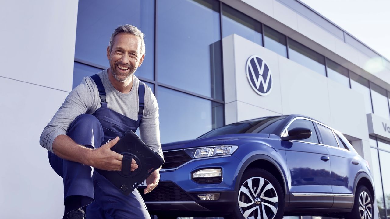 Ein Volkswagen Service Techniker mit Tablet, kniet vor einem blauen Volkswagen T-Roc