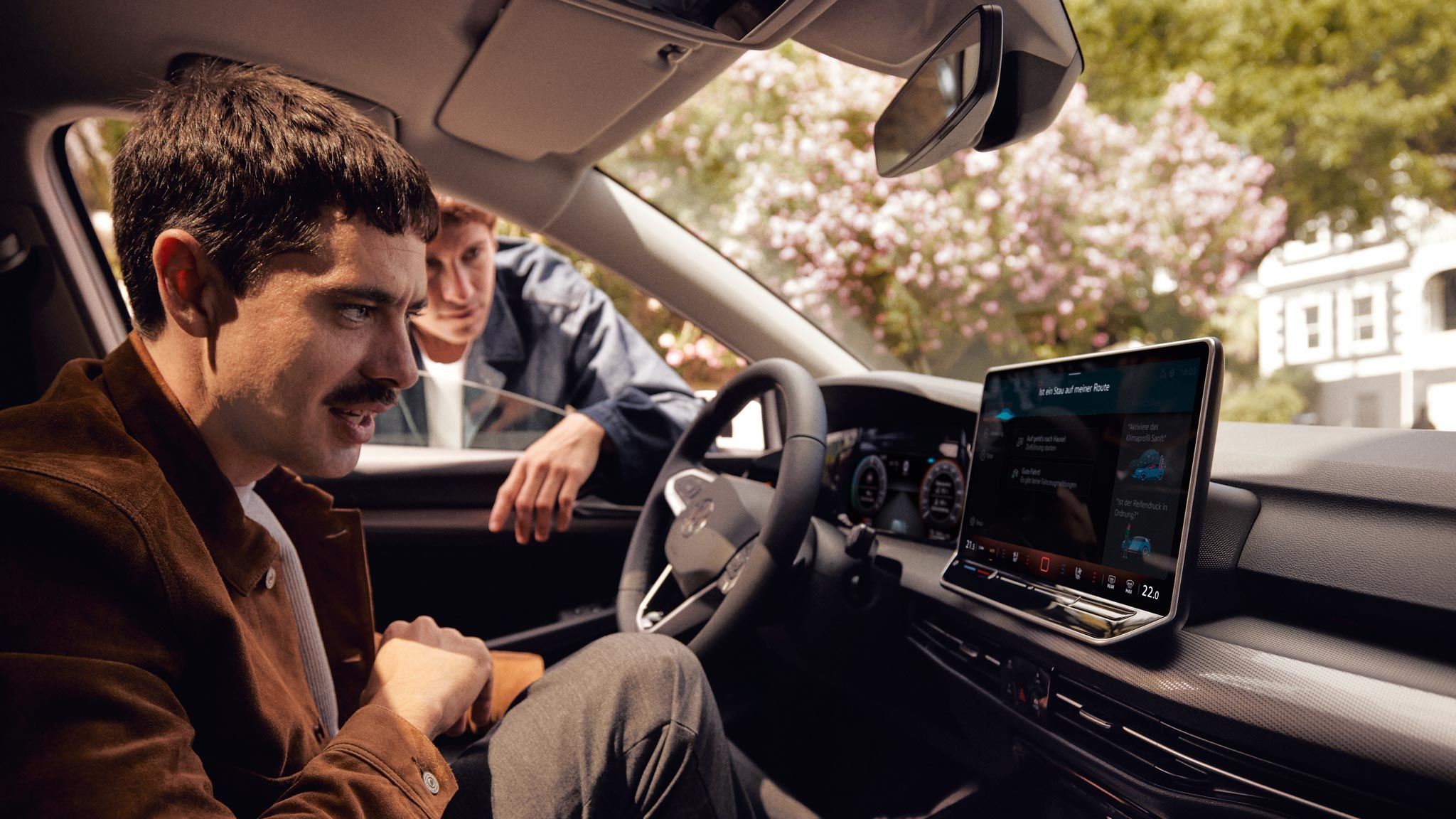 Ein Mann sitzt im neuen VW Golf und schaut auf das Infotainment-Display, ein anderer schaut durch das Fenster.