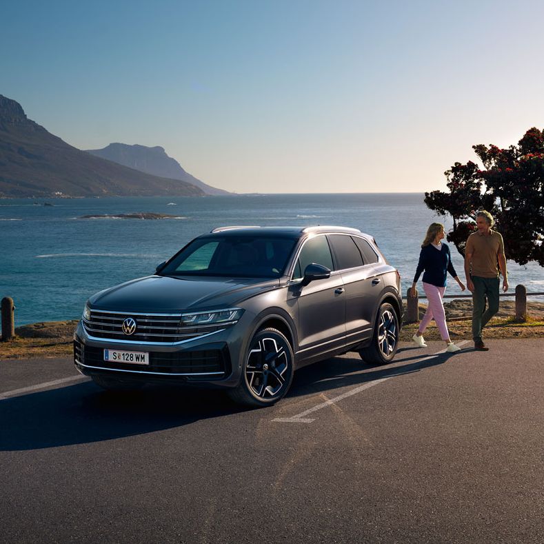 Der neue Volkswagen Touareg auf einem Parkplatz an der Küste, an ihm gehen eine Frau und ein Mann vorbei