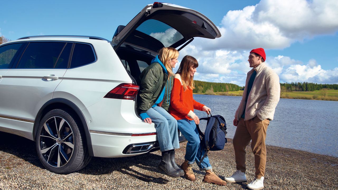 Zwei Frauen sitzen am Kofferraumeinstieg des VW Tiguan Allspace, ein Mann steht daneben